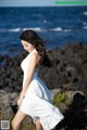 A woman in a white dress standing on a rock by the ocean.