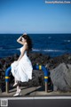 A woman in a white dress standing on a railing by the ocean.