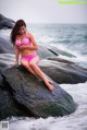 A woman in a pink bikini sitting on a rock by the ocean.