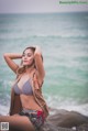 A woman in a bikini sitting on a rock by the ocean.