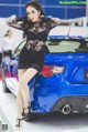 A woman in a black dress standing next to a blue car.